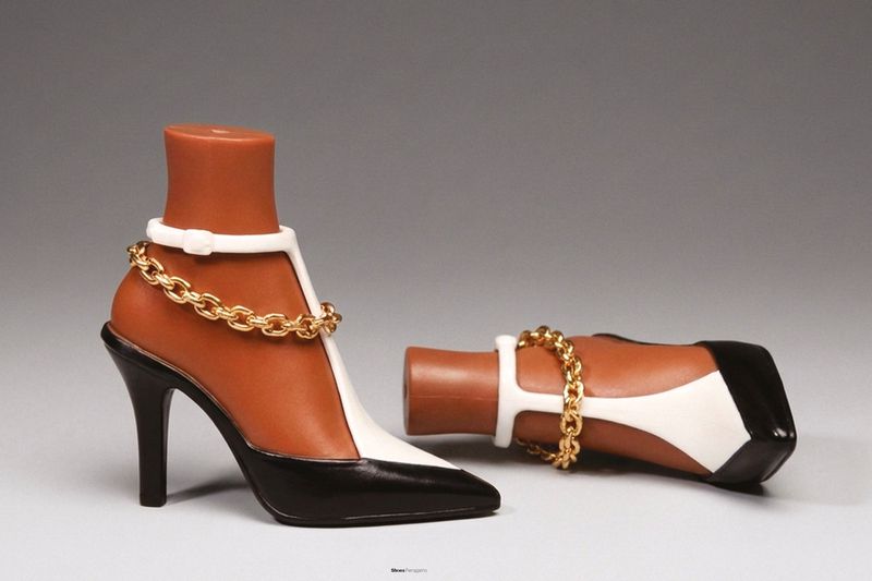 A pair of brown and black high heels with white accents and gold chain detail are displayed on a light surface against a grey backdrop.