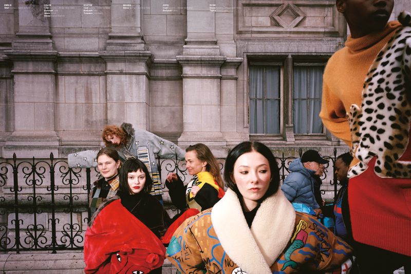 Fashion models in colorful graphic coats and shearling jackets posed along an ornate black iron railing outside a stone building.