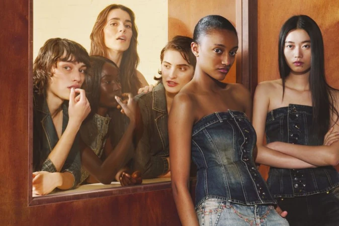 Group of six models in denim corsets and jackets posing around a warm wood-paneled partition.