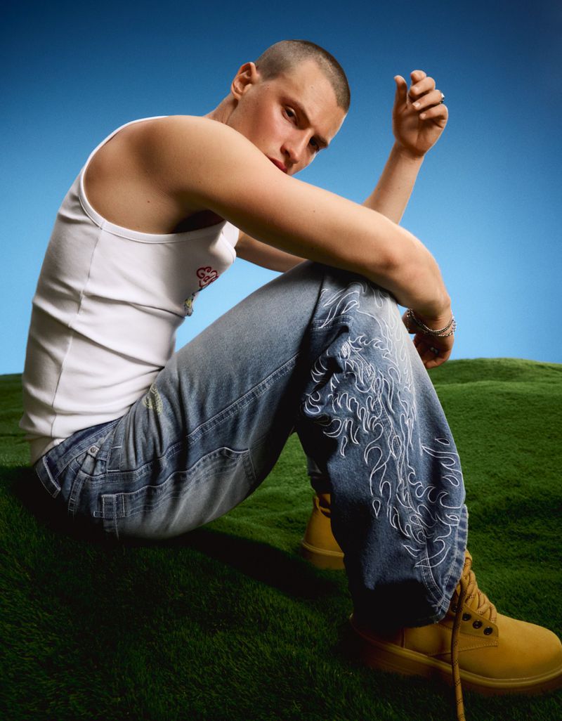 Man with a buzz cut wearing a white tank top and flame-embroidered blue jeans seated on green grass against a blue backdrop.