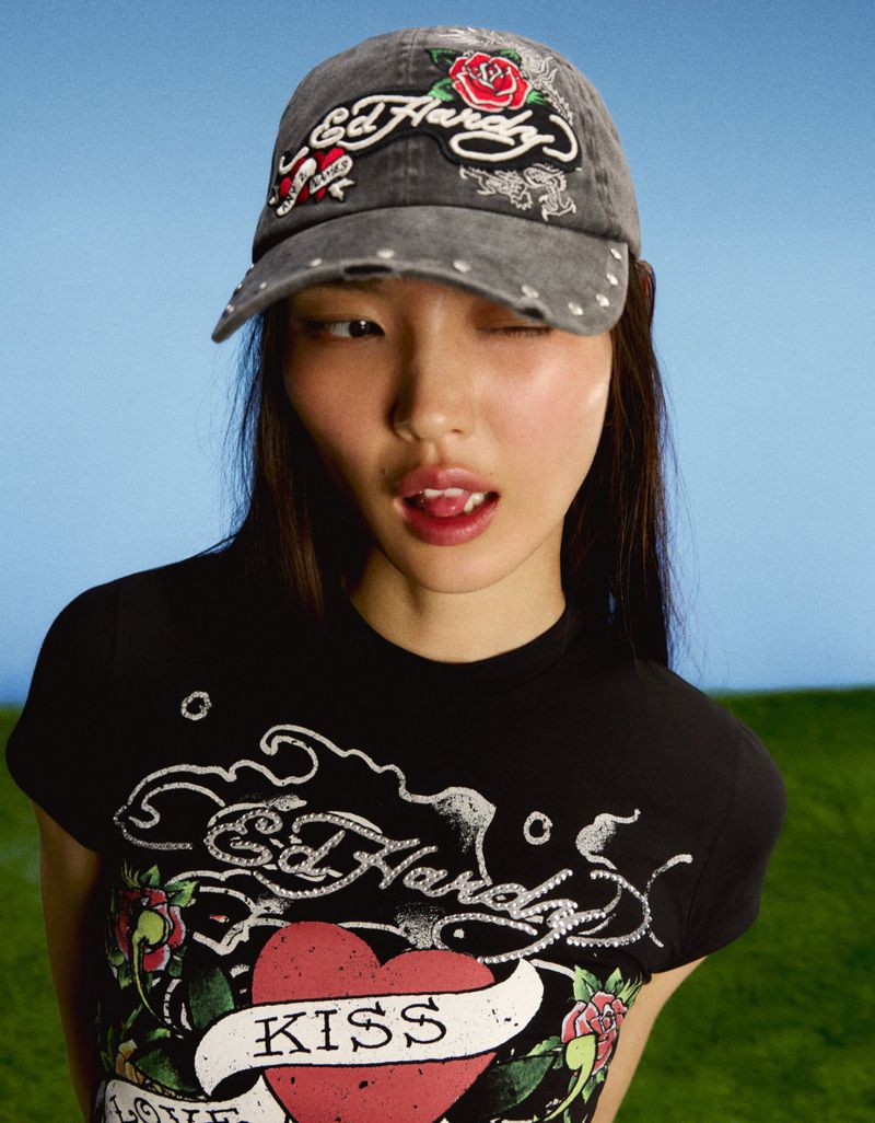 Model Lian Jia wearing a washed grey Ed Hardy embroidered baseball cap and black graphic t-shirt against a blue and green background.