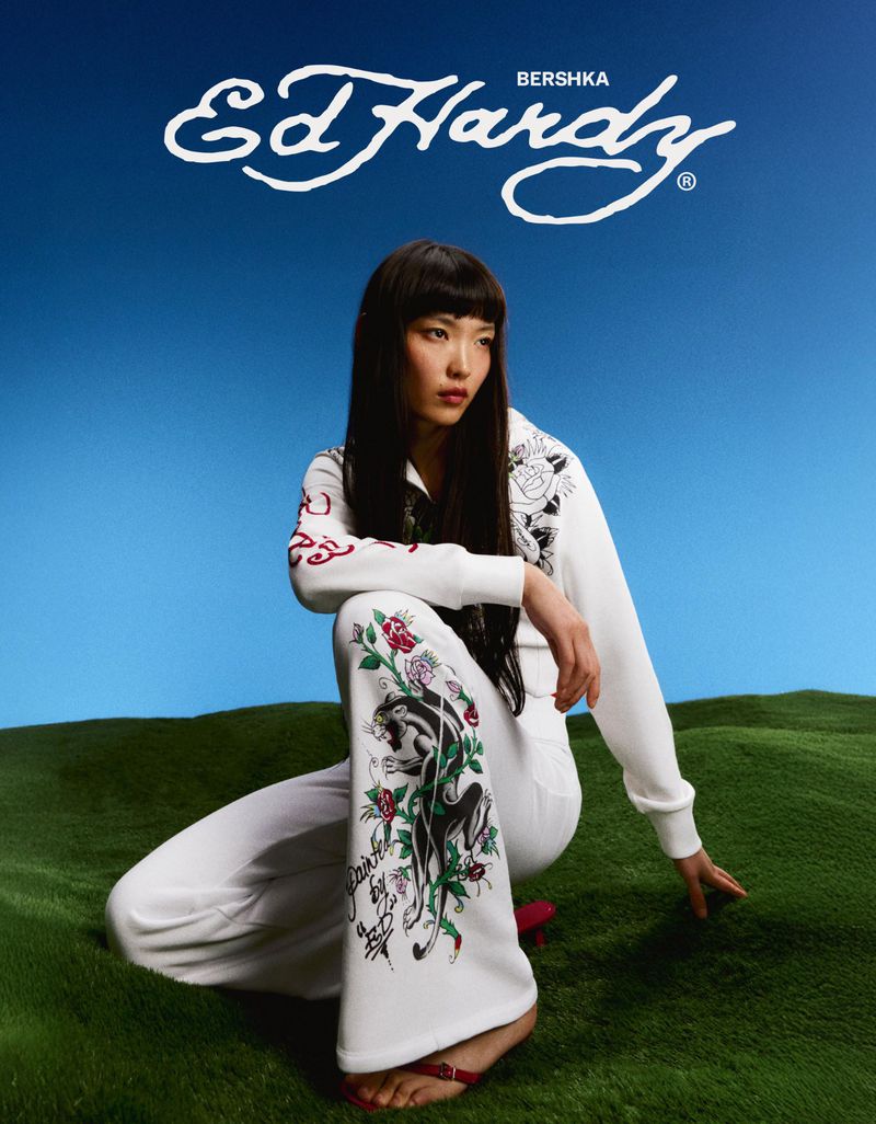 Model Lian Jia wearing a white Ed Hardy graphic tracksuit while crouching on green grass against a clear blue sky.