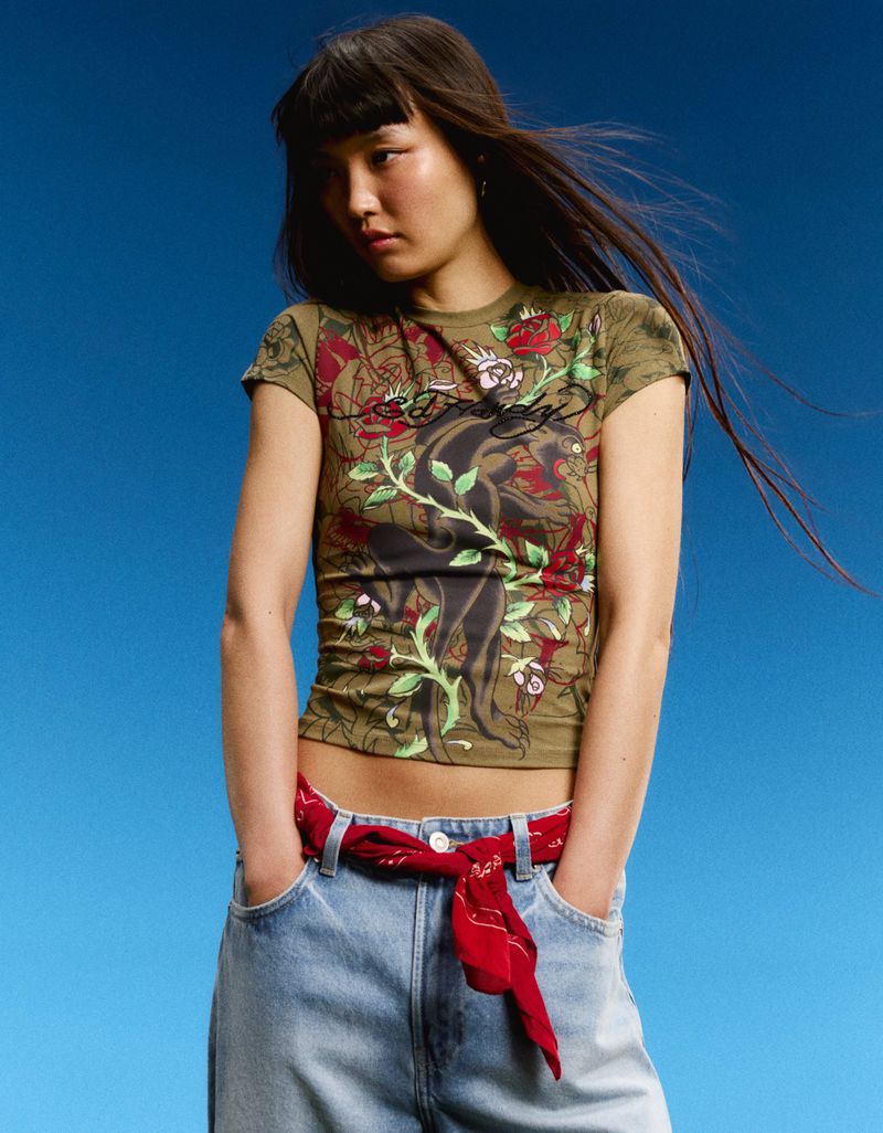 Model Lian Jia wearing an olive green Ed Hardy graphic baby tee, light wash jeans, and a red bandana belt against a blue backdrop.
