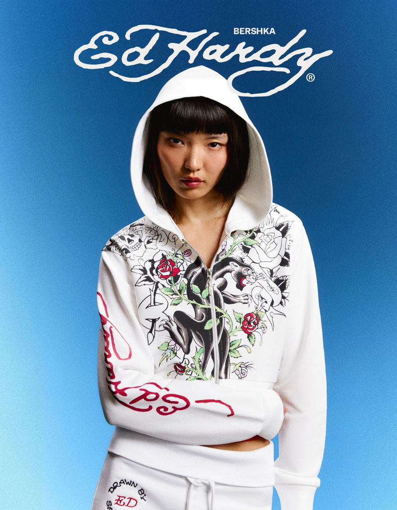 Model wearing a white Ed Hardy zip-up hoodie with graphic panther and rose prints against a blue gradient backdrop