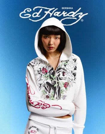 Model wearing a white Ed Hardy zip-up hoodie with graphic panther and rose prints against a blue gradient backdrop