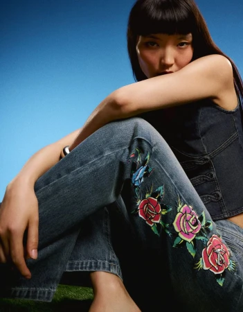 Model Lian Jia wearing floral embroidered denim jeans and a dark denim top, seated outdoors against a clear blue sky.