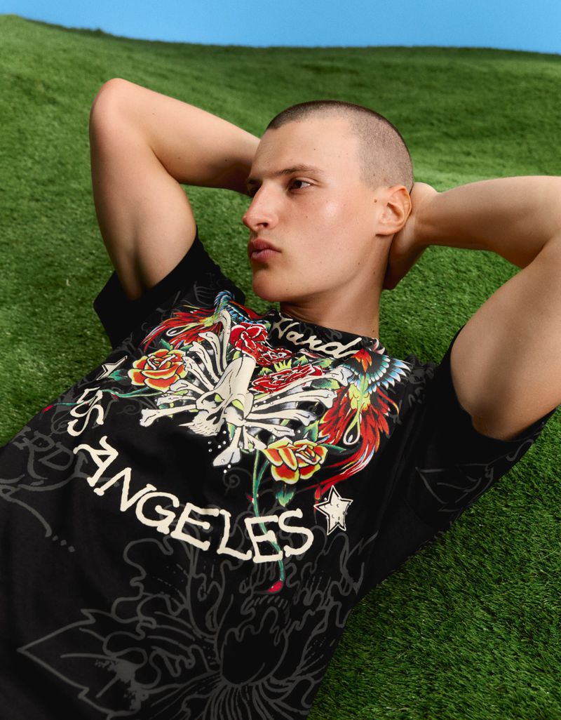 Man with buzz cut lying on green grass with hands behind head wearing black Ed Hardy graphic t-shirt.