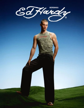 Male model in an olive graphic tank top and black wide-leg trousers posing on a grassy mound against a blue sky backdrop.