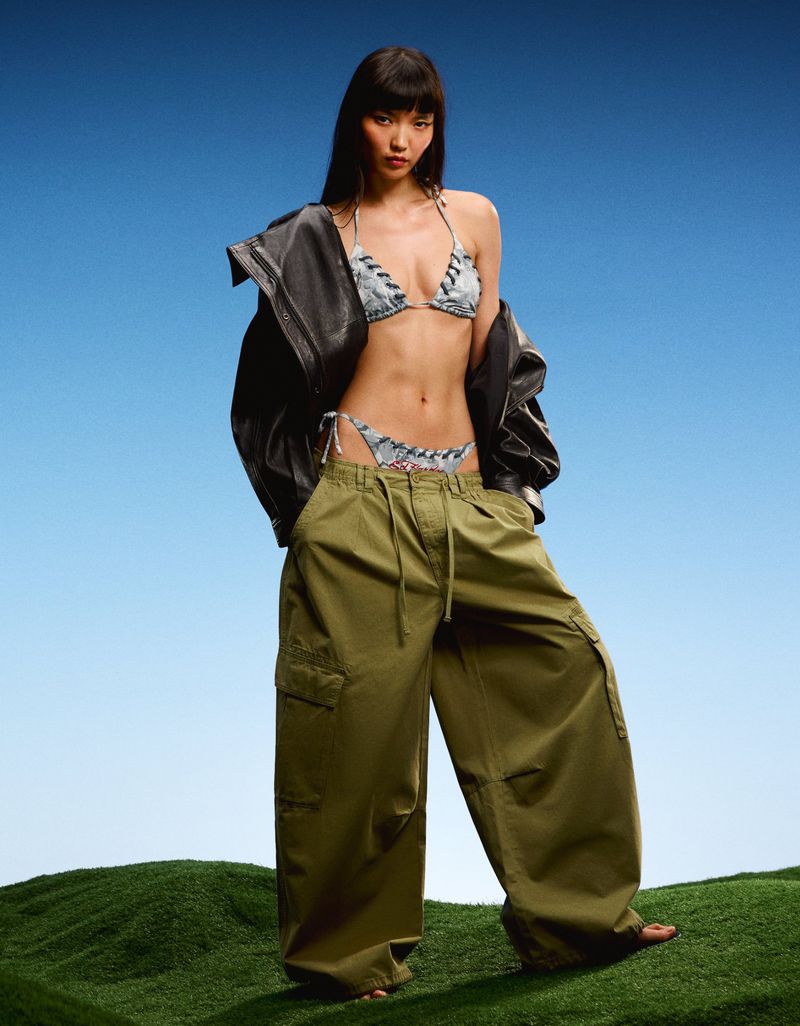 Model Lian Jia wearing an oversized black leather jacket, printed bikini, and wide-leg olive cargo pants on grass against a blue backdrop.