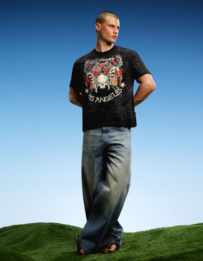Model wearing a black Ed Hardy graphic t-shirt and wide-leg denim jeans standing on a grassy hill against a blue sky.
