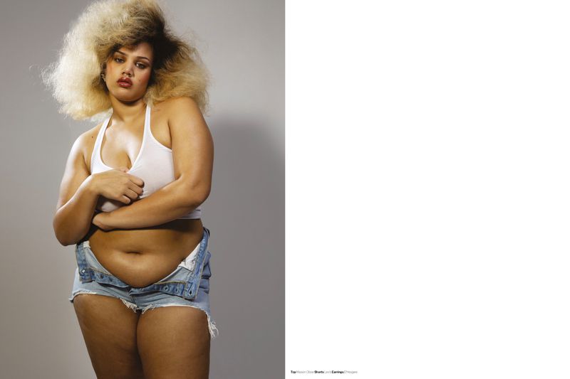 Portrait of a woman with curly blonde hair, wearing a white tank top and denim shorts, posing in front of a grey backdrop.