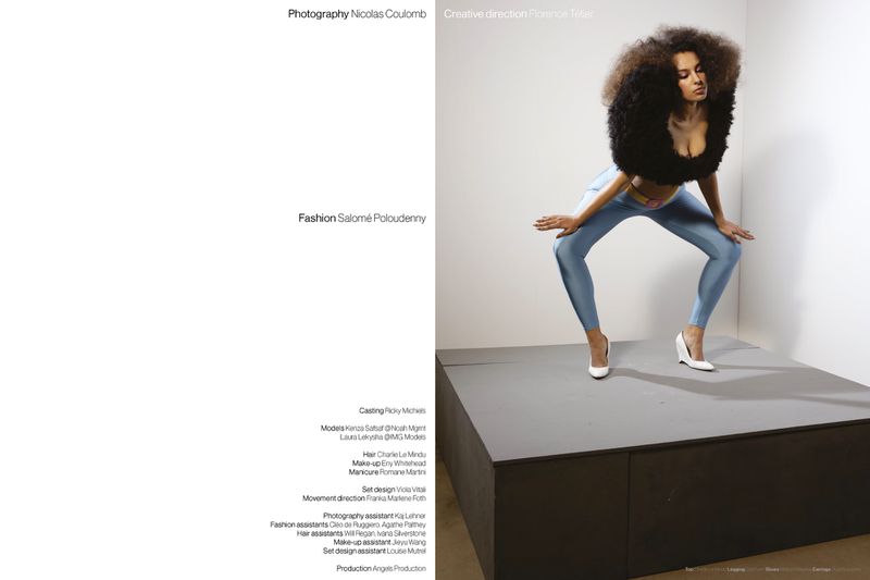 Fashion model in a voluminous black textured shrug, blue satin leggings, and white heels posing dynamically on a grey platform.