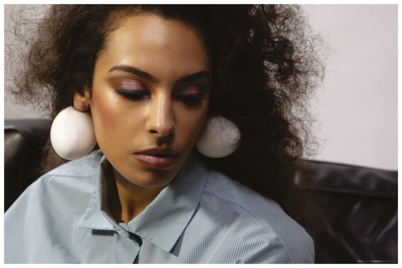 Close-up of a woman with voluminous curly hair wearing large egg earrings and a light blue collared shirt.