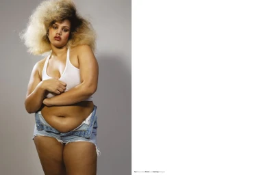 Model with voluminous blonde hair wearing a white crop top and unbuttoned denim shorts against a neutral grey studio background