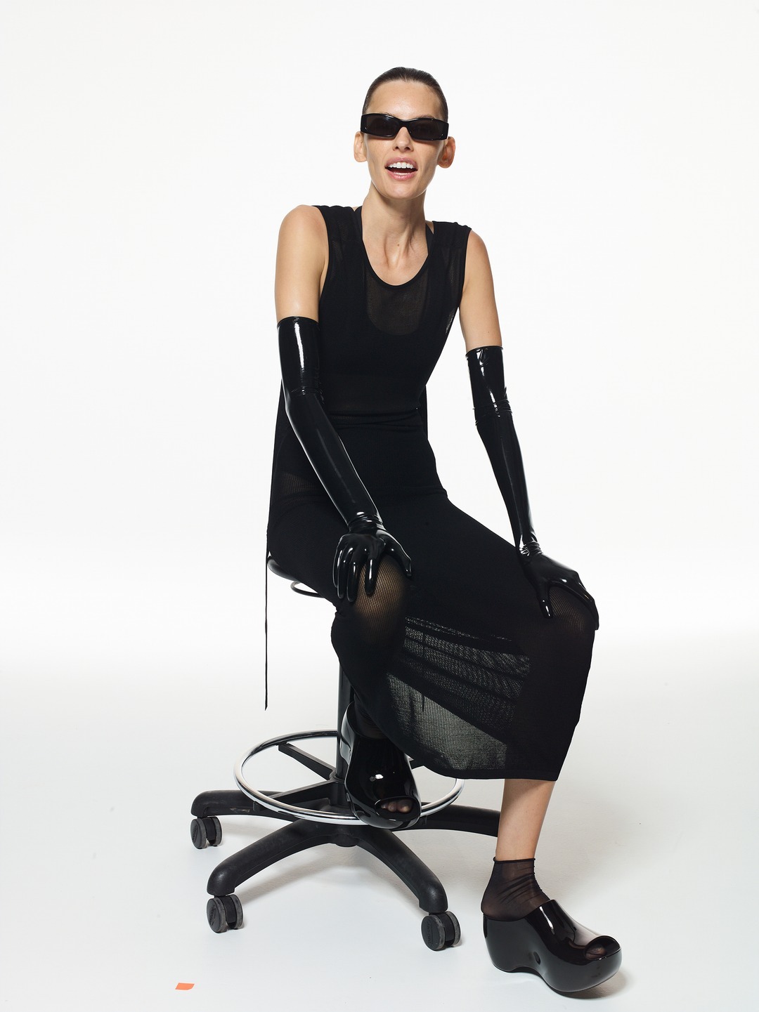 Adult woman in a black dress sits on a rolling stool in front of a white background, wearing black sunglasses and shiny black gloves.