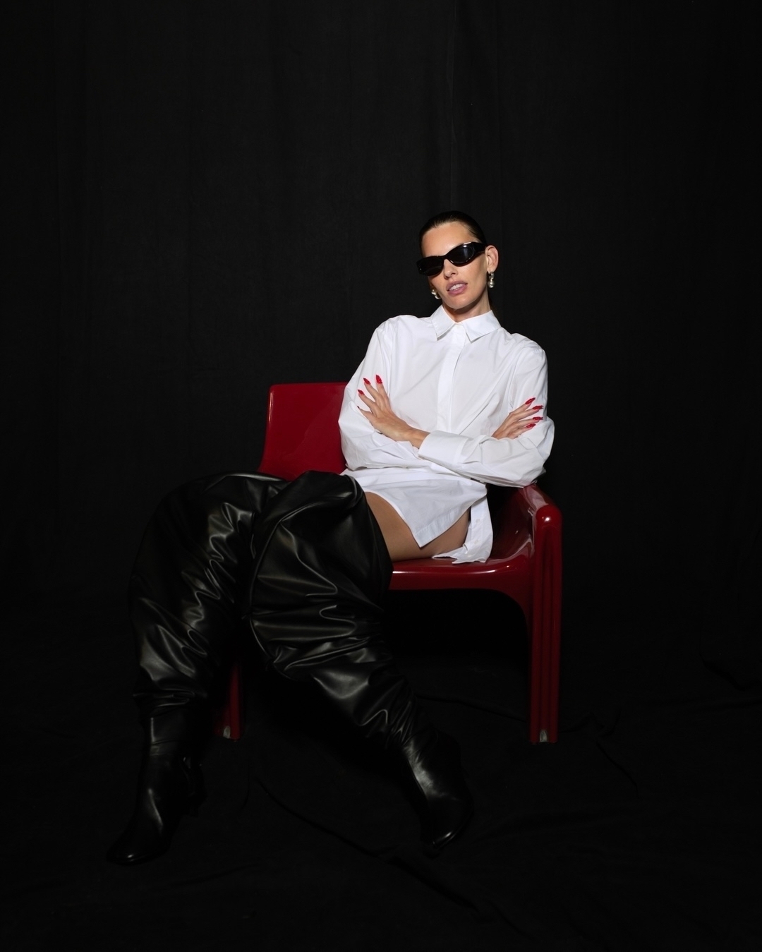Model wearing dark sunglasses, white shirt, and black boots sitting on a red chair against a black backdrop