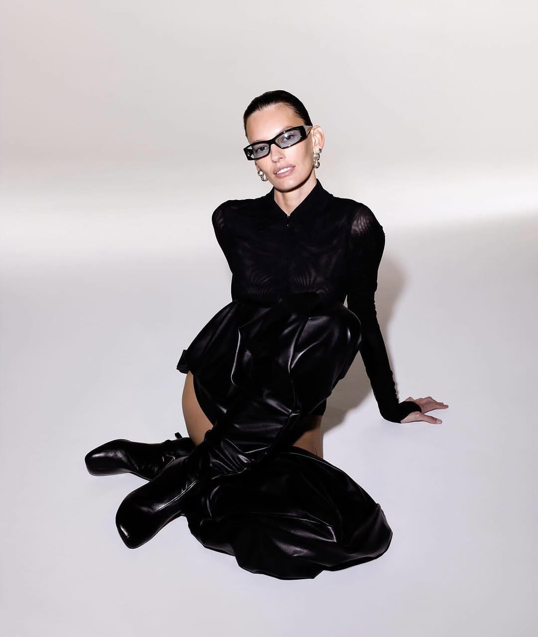 Elegant adult woman in black top, dark glasses, and black leather over-the-knee boots poses against a bright gradient background.