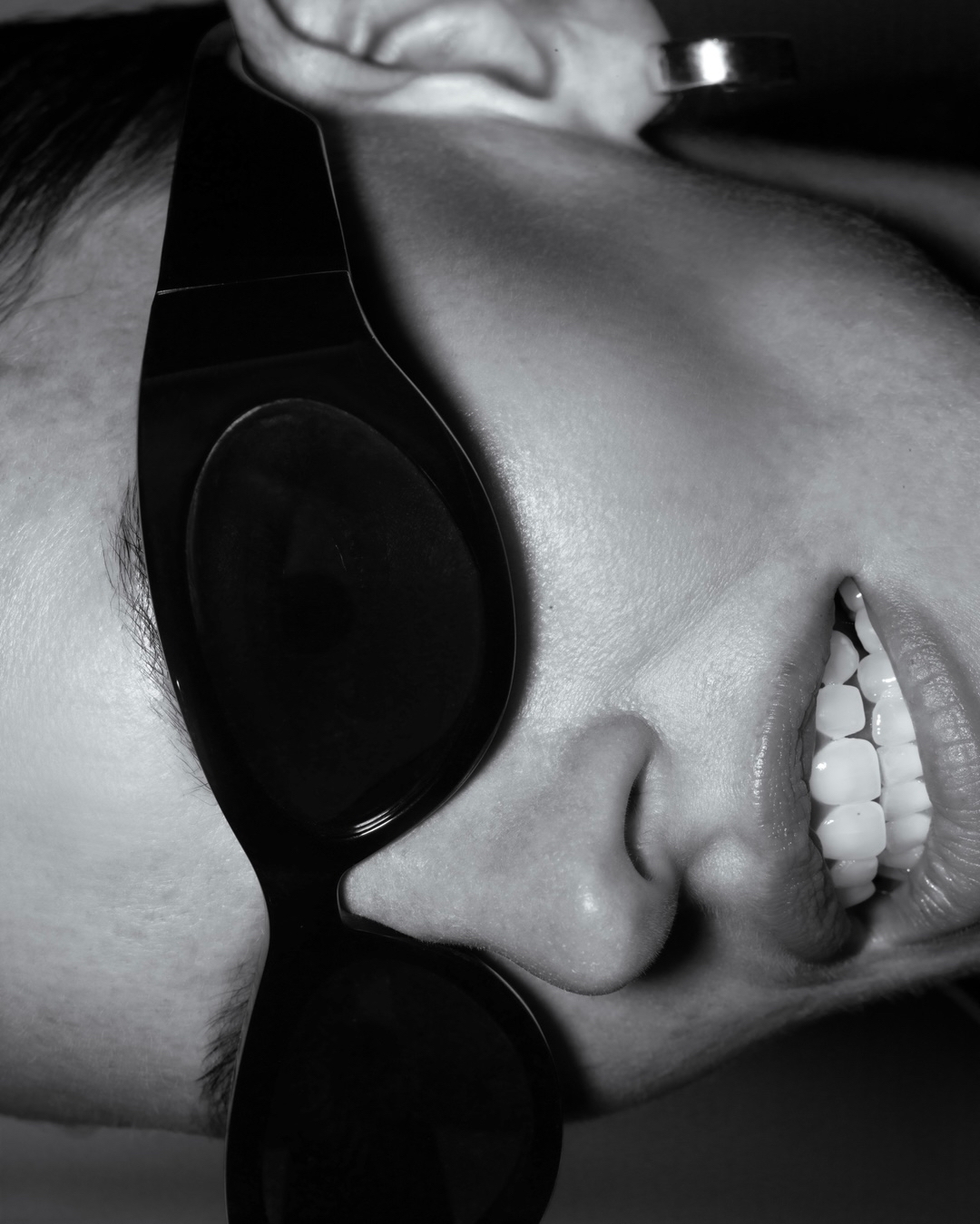 Close-up portrait of a model wearing thick black sunglasses with visible teeth and a silver earring in a high-contrast monochrome framing.