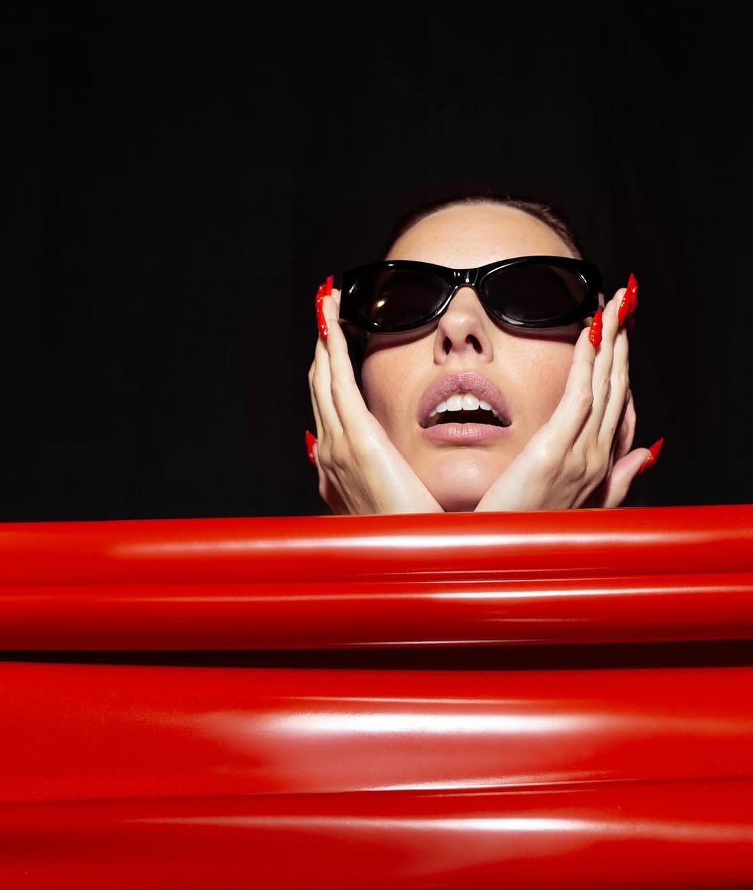Woman with red nails and sunglasses poses behind a red tubular structure against a dark background in a beauty fashion portrait.