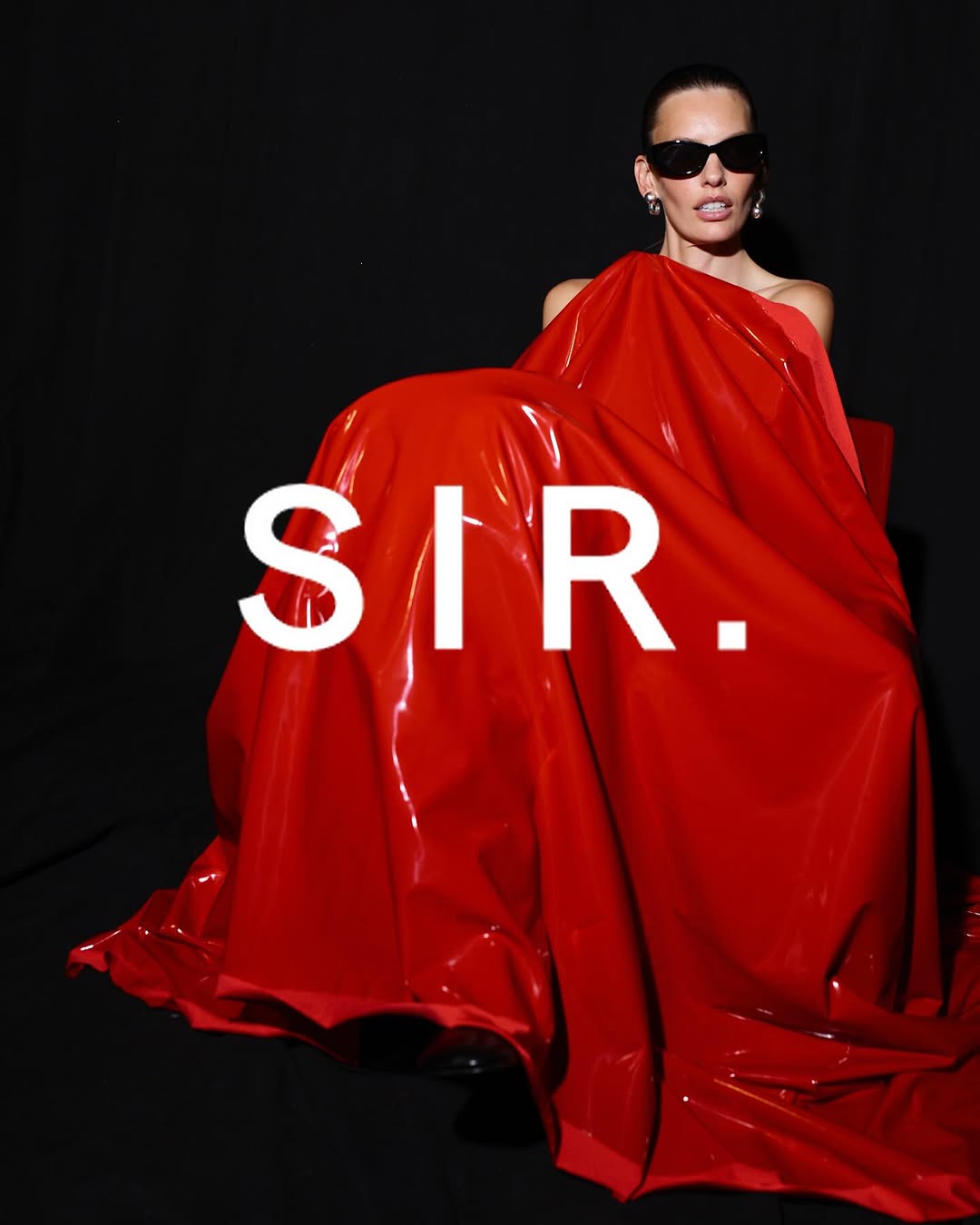 Glamorous woman with sunglasses draped in glossy red fabric poses in a dark studio. Fashion and portrait photography.