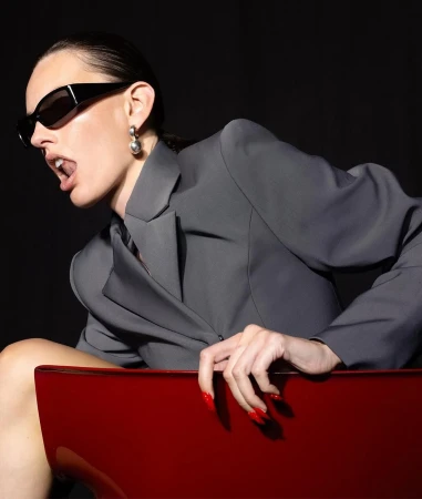 Woman in a grey blazer poses with an open mouth and dark sunglasses. She sits on a red chair against a dark studio backdrop.