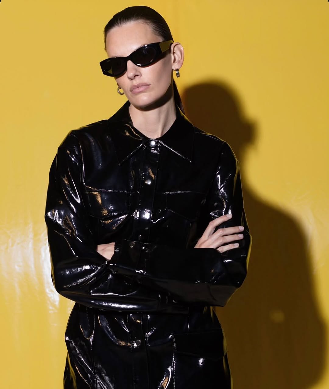 Stylish adult woman in sunglasses and black patent leather jacket with arms crossed against a bright yellow studio backdrop.