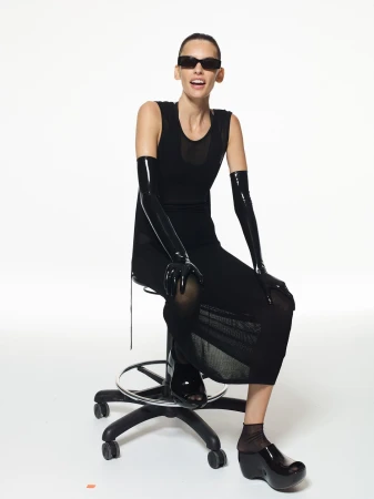 Model seated on a rolling stool wearing a black dress, black sunglasses, and long shiny black gloves against a white background