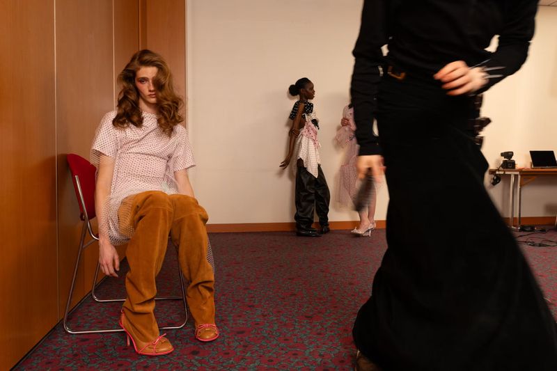 Model in wide-leg caramel trousers and sheer patterned tunic sits on a chair in a backstage room while others stand in the background.
