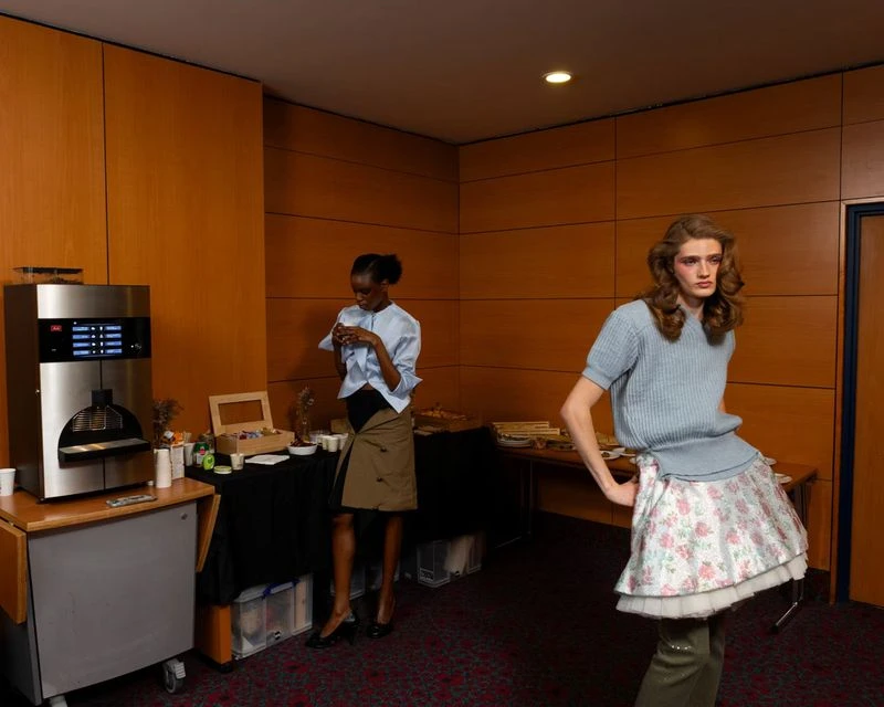 Two women pose in an indoor setting with a coffee machine and buffet table. One woman wears a blue top and khaki skirt, another woman wears a blue top and floral skirt.