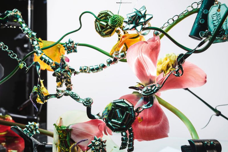 Sculptural metallic green bow jewelry and ribbons intertwined with pink tulip petals and fresh floral stems against a white backdrop.