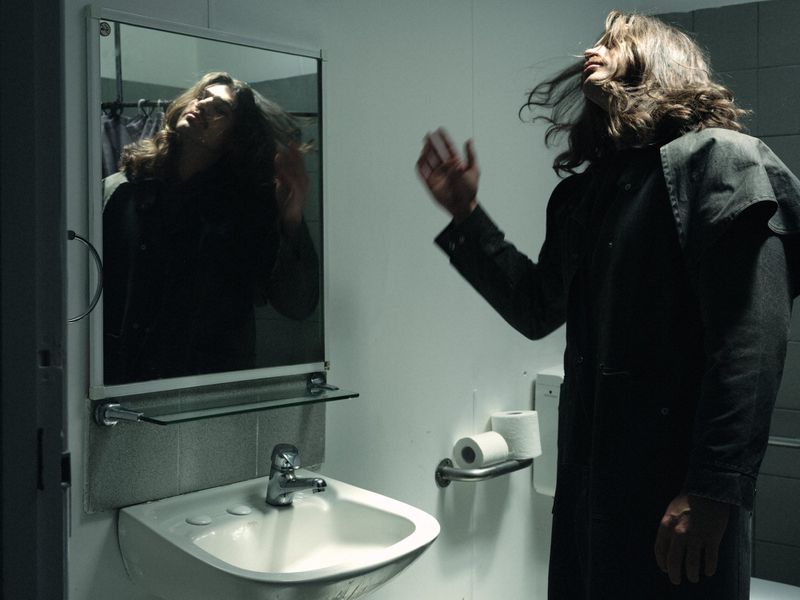Long-haired man in a dark trench coat tossing his hair in front of a bathroom mirror and porcelain sink.