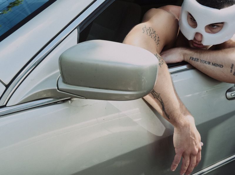 Shirtless man wearing a white mask leaning out of a silver car window with tattoos visible on his arms