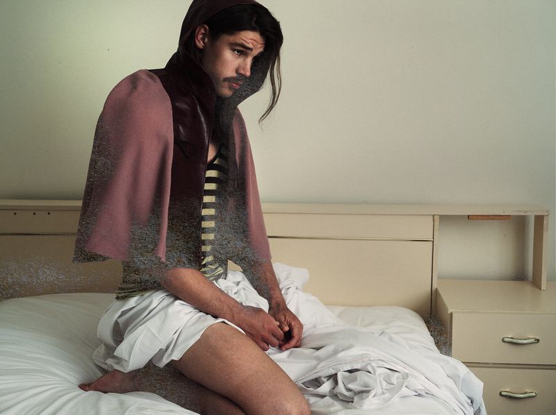 Man with a mustache wearing a hooded cape and shorts kneeling on an unmade bed with a digital sand dissolve effect across his torso.