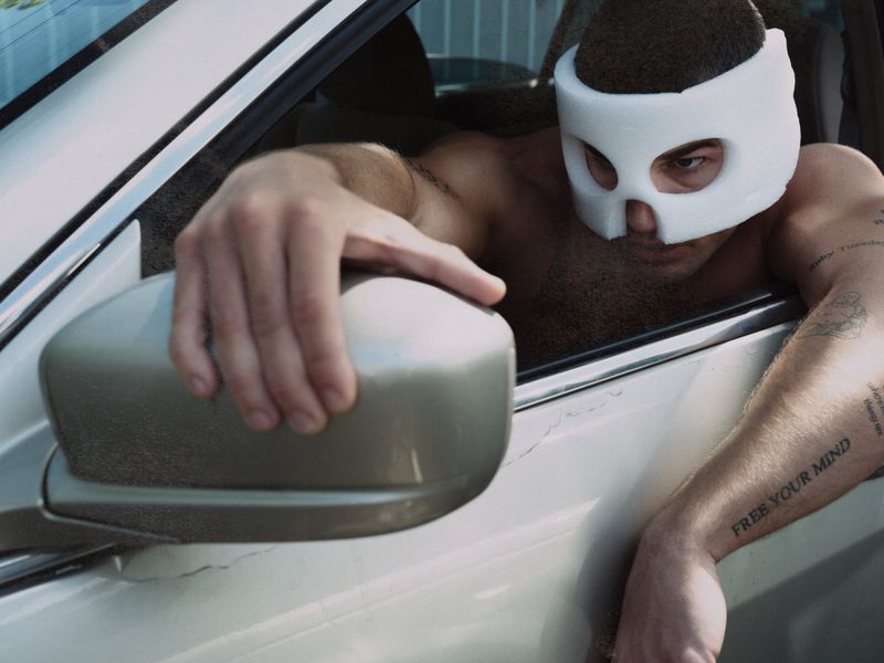 A shirtless man wearing a white padded face mask leans out of a car window, gripping the side mirror with visible arm tattoos.