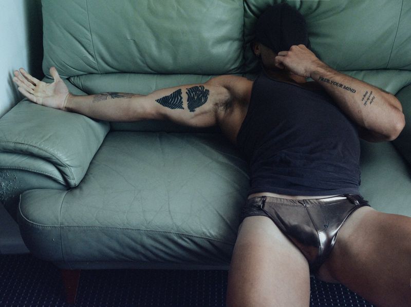 A man in a black tank top and metallic briefs lying back on a green leather couch with his face obscured by a cap.