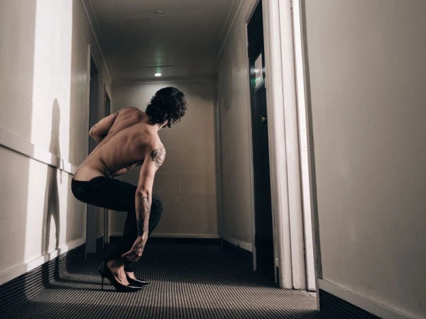 Shirtless man with tattoos crouching in a narrow hallway wearing black trousers and high heels