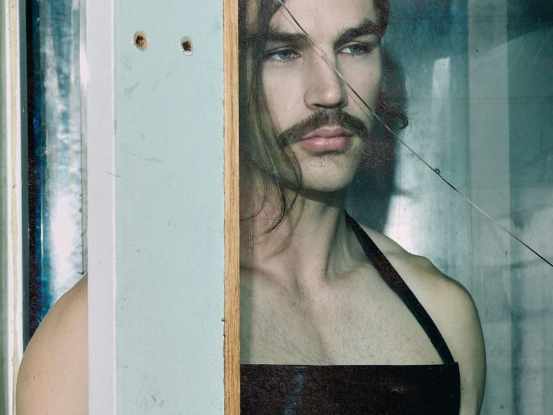 Man with a mustache and long brown hair wearing a black halter strap top, seen through a cracked glass frame.