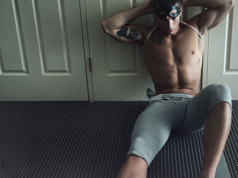 Shirtless man wearing a black mask and grey cropped sweatpants seated on a patterned floor with hands behind his head.