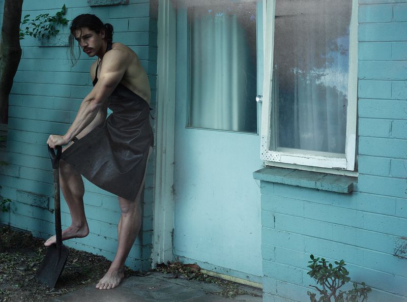 Man in a dark apron holding a shovel while standing barefoot against a light turquoise exterior wall.