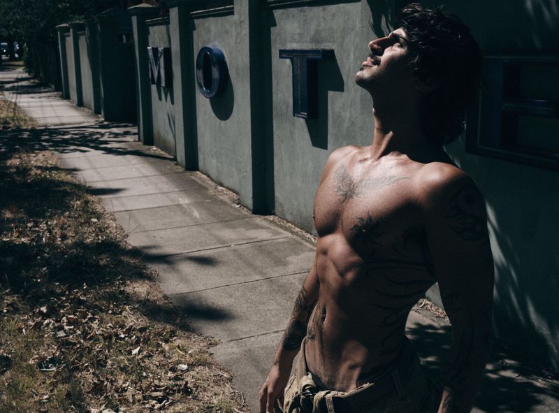 Shirtless man with tattoos leaning back on a sidewalk beside a motel sign wall in harsh sunlight.