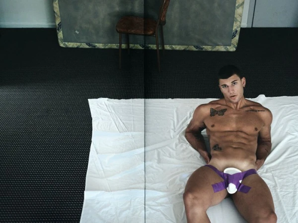 Muscular shirtless man reclining on a white sheet laid across dark carpet, wearing a protective athletic cup taped with purple adhesive.