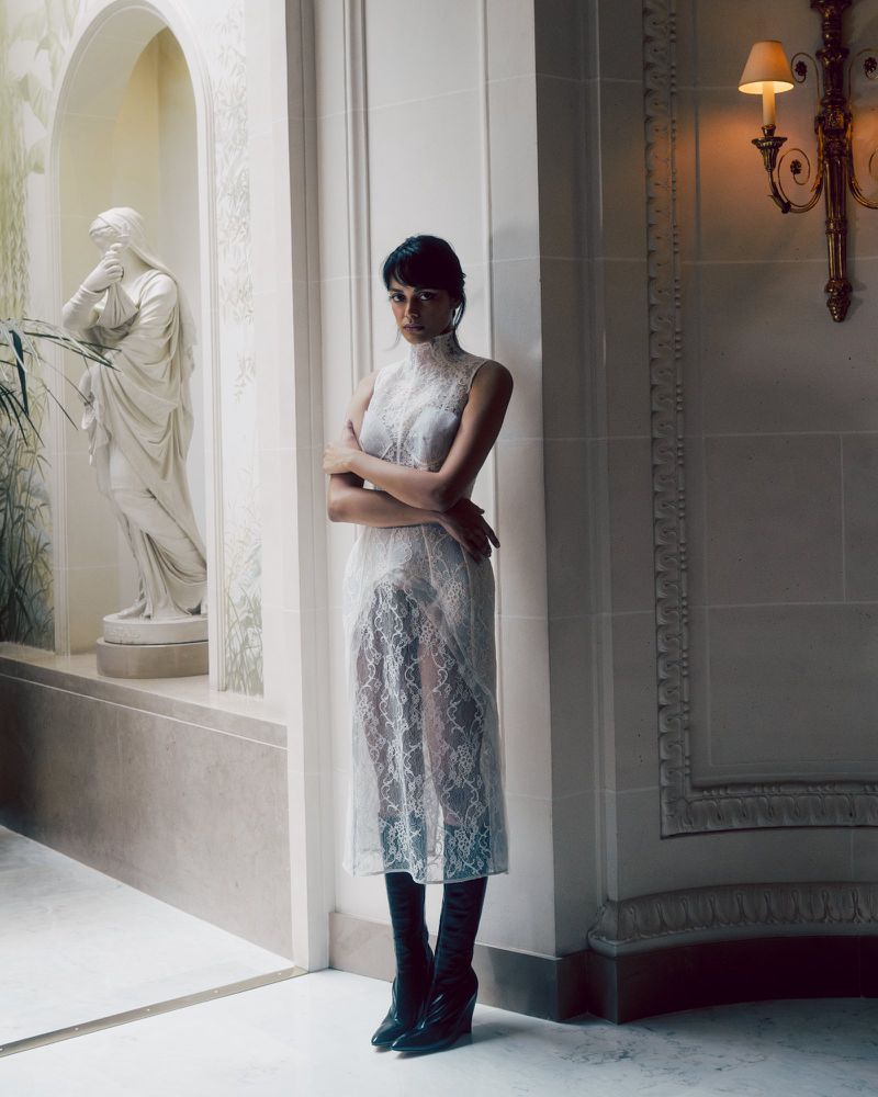 Model wearing a sheer lace dress and dark boots standing in a classical interior with a marble statue.