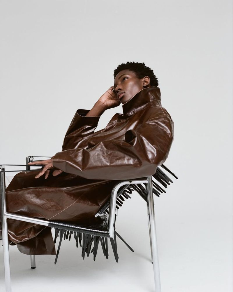Model Ibra Niakate reclines in a chrome armchair wearing an oversized brown leather coat against a white studio background.