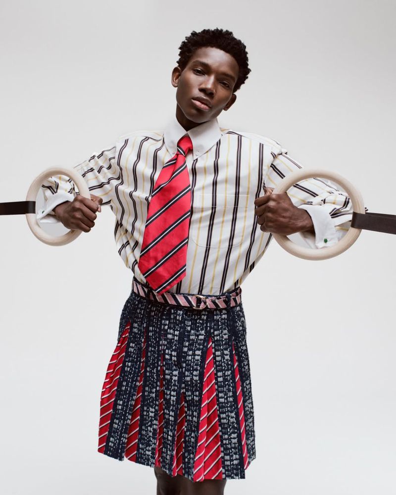Model Ibra Niakate holds wooden gymnastic rings wearing a striped shirt, red tie, and pleated kilt against a white studio backdrop.