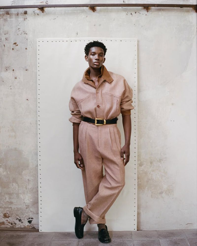 Model Ibra Niakate wearing a dusty rose collared jumpsuit with a black belt and loafers against a studio panel for Esquire Mexico.