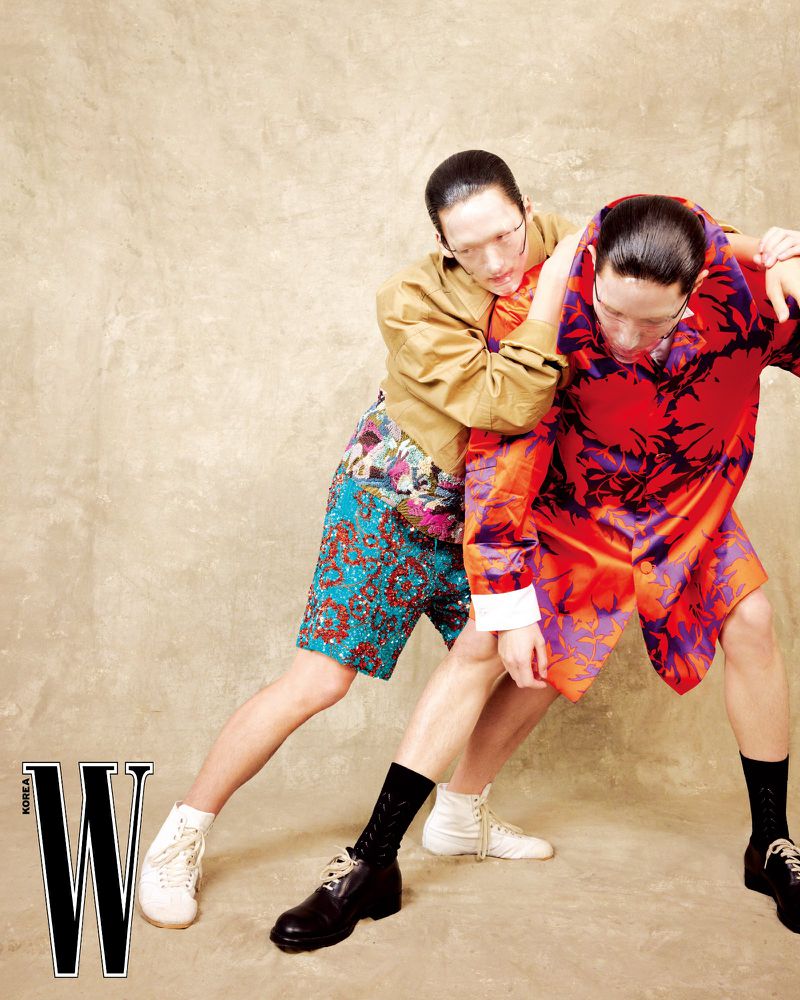 Two models in vibrant patterned clothing posing dynamically against a textured neutral wall for W Korea fashion editorial.