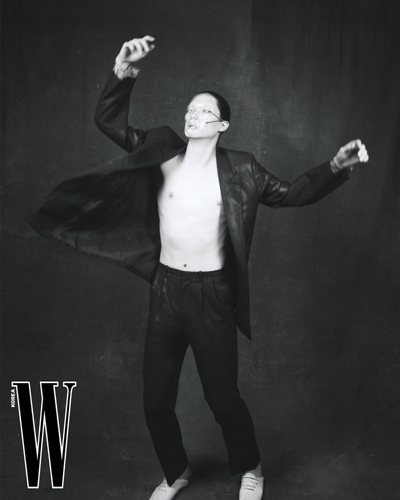 A person posing in a black jacket and pants against a dark background, with a 'W Korea' logo visible on the left side of the frame.