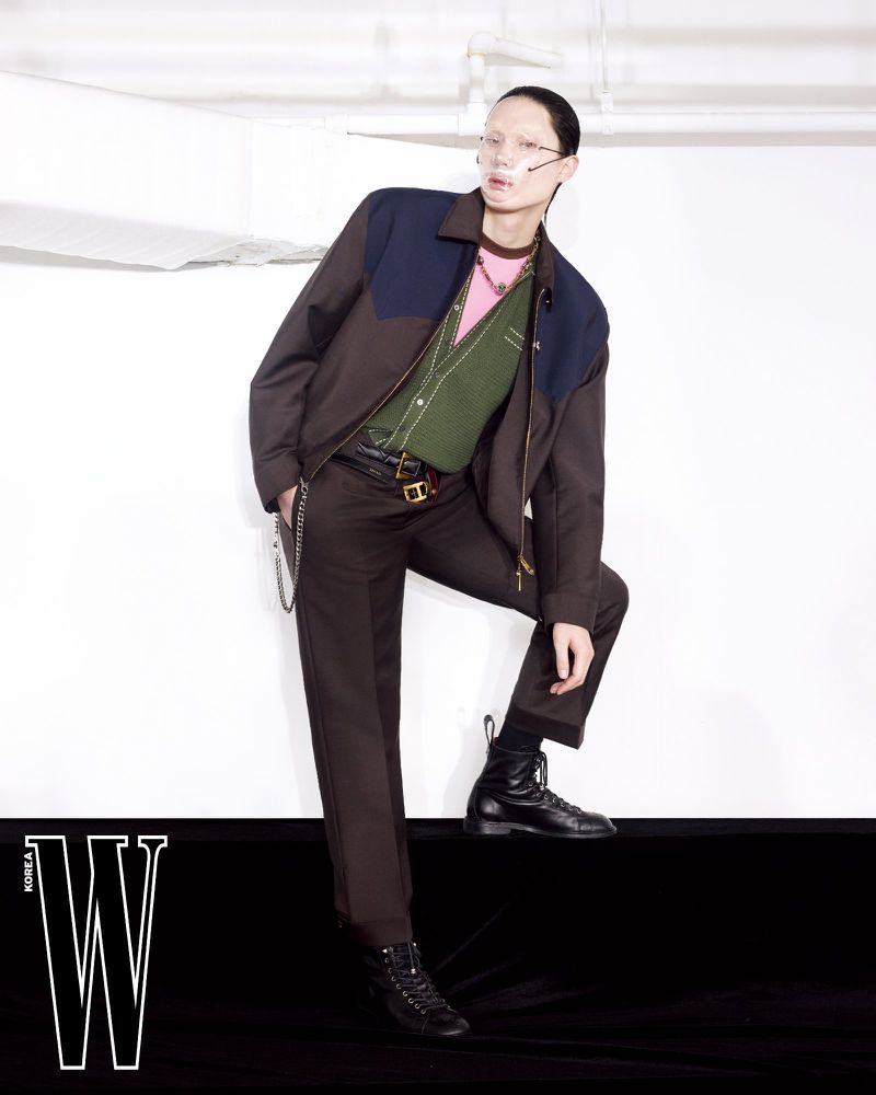 Model wearing a brown jacket over a green vest and trousers standing against a plain white wall