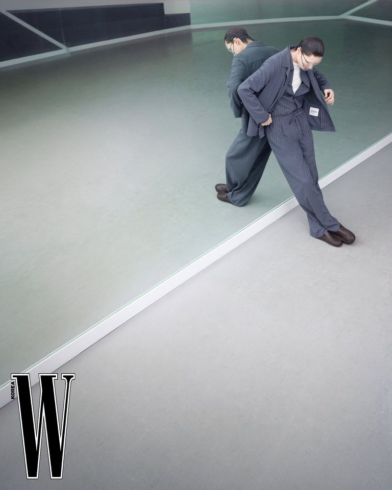 Model Jinyeong Pak wearing a pinstripe suit leaning against a large mirror in a minimalist studio setting.