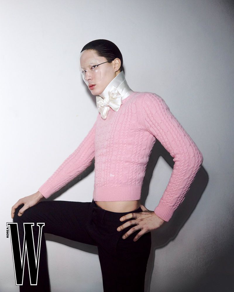 Model Jinyeong Pak wearing a cropped pink cable-knit sweater, white satin bow collar, and black trousers against a light wall.